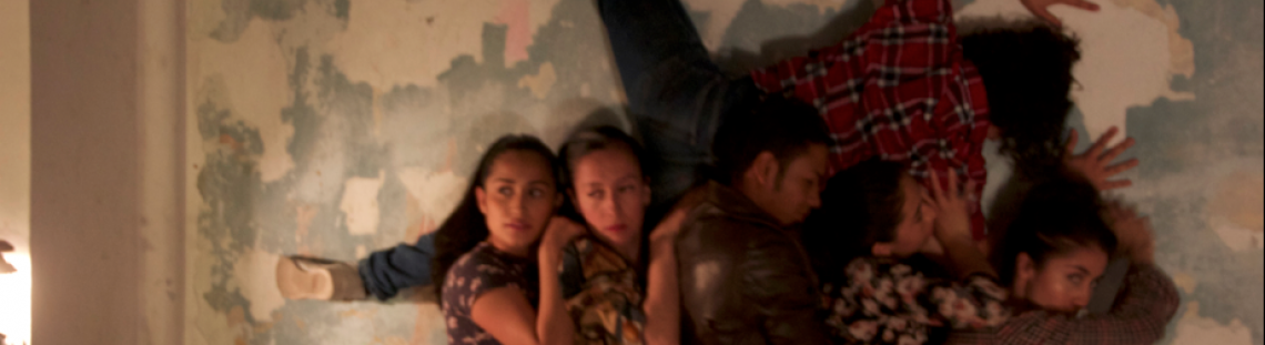 Actores sosteniendo en alto un cuerpo contra una pared.