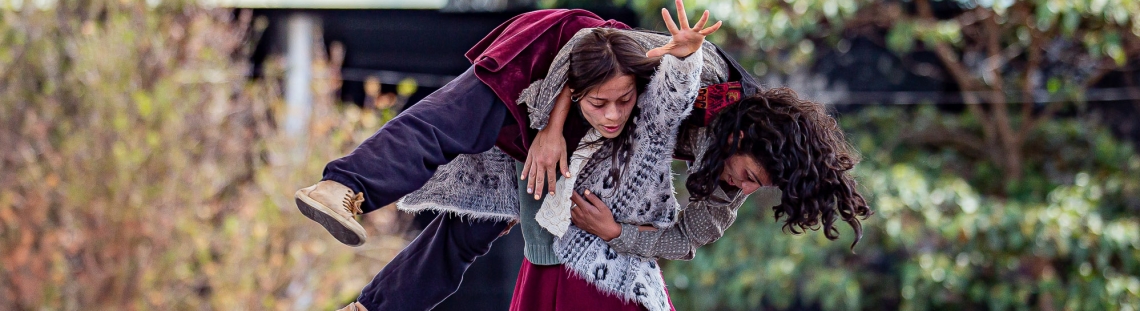 Dos mujeres bailan en tarima, una alza a la otra en su espalda. 