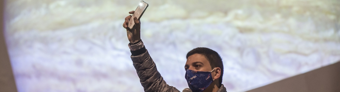 Hombre levantando la mano con un celular con una proyección de Júpiter de fondo. 