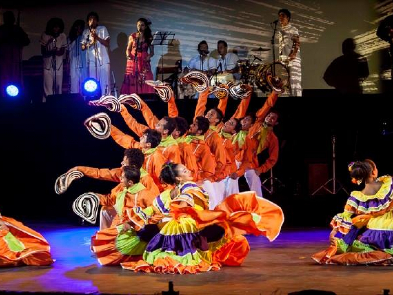 Grupos debailarines vestidos de colores en escenario con grupo musical al fondo.