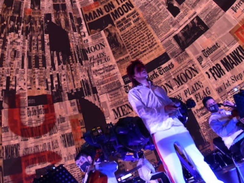 Hombre vestido como David Bowie en la tarima del domo del Planetario con imágenes de periódicos en el fondo. 