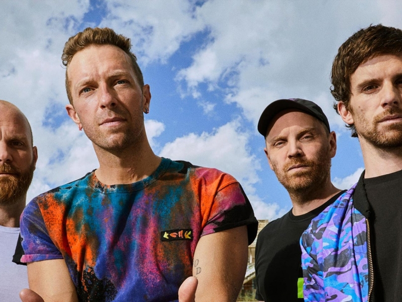 Integrantes de Coldplay con cielo azul y nubes de fondo. 