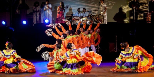 Grupos debailarines vestidos de colores en escenario con grupo musical al fondo.