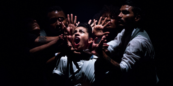 Varios hombres en el escenario, uno de ellos en actitud de grito rodeado por las manos de los otros.