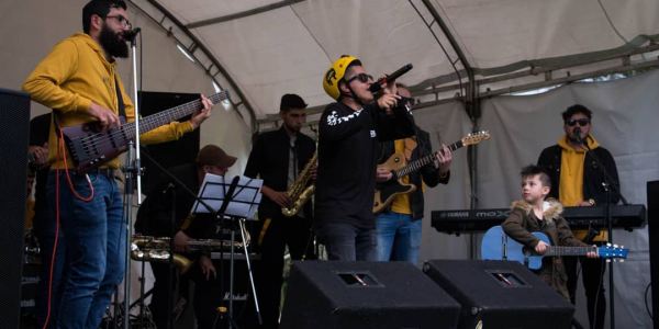 Agrupación musical realizando una presentación en vivo sobre una tarima en espacio abierto, en la cual se ven siete hombres adultos, y un niño sosteniendo una guitarra azul.