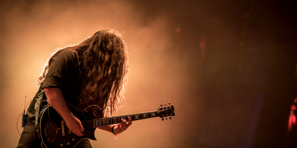 Hombre de pelo largo tocando una guitarra eléctrica con una luz amarilla de fondo, en un evento musical