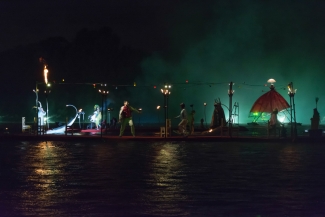 Actores en el muelle sobre el lago, con sillas, luces y faroles.