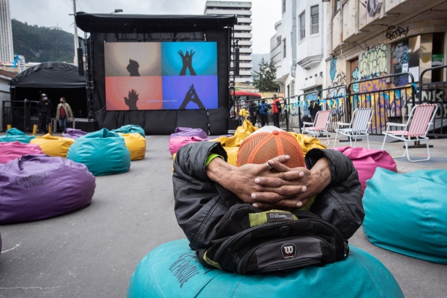 Fotografía de señor sentado en puff con las manos cruzadas detrás de la cabeza viendo una proyección audiovisual 