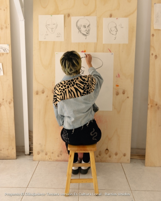 Fotografía de persona sentada en una butaca dibujando a mano alzada sobre papel colgado en pared.